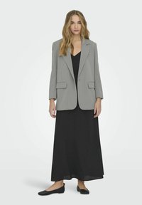 Blazer gris a rayas con grandes solapas, dos bolsillos delanteros y cierre de un solo botón, llevado sobre un vestido maxi negro y zapatos planos negros.