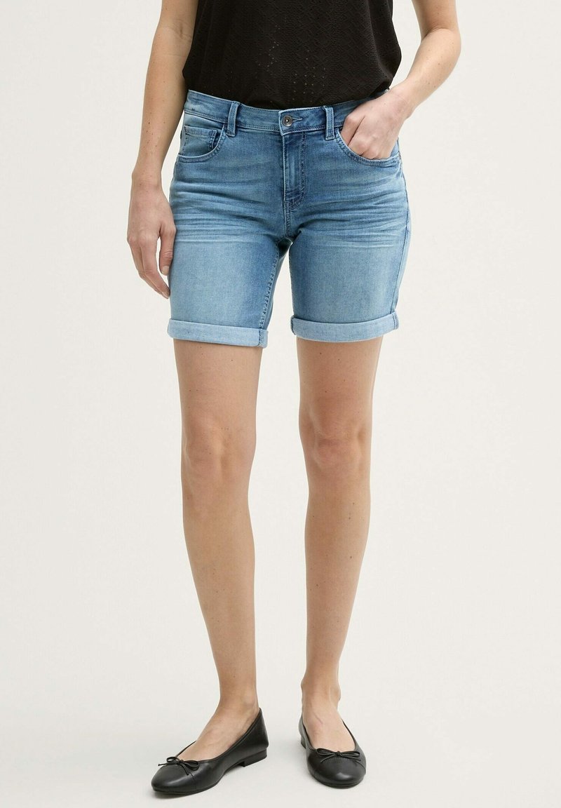 Pantalones cortos de mezclilla en azul claro con un corte clásico, dobladillo enrollado y un desvanecido sutil. Combinados con zapatos planos negros y una blusa de manga corta.