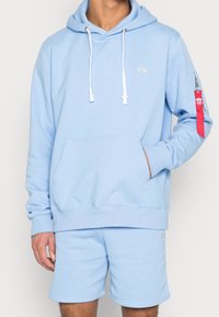 Felpa azzurra chiara con tasca frontale, coulisse bianche e un'accenzione rossa sul maniche. Abbinata a pantaloni corti azzurri chiari.
