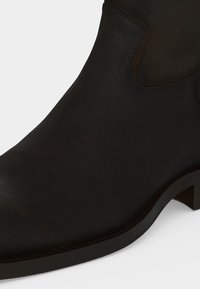 Gros plan d'une bottine en cuir noir avec un petit talon et un bout arrondi lisse sur fond blanc.
