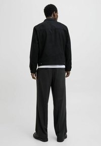 Sort ruskindsjakke med krave og knapper, kombineret med grå plaidbukser. Outfittet har en afslappet pasform og en glat tekstur.