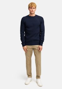 Marineblaues, geripptes Strickpullover mit langen Ärmeln, Rundhalsausschnitt und eng anliegenden Bündchen, kombiniert mit beigen Hosen und weißen Sneakers.