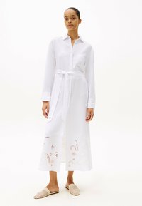 Vestido camisa blanco de tela ligera, con cintura ceñida con cinturón, tapeta de botones y bordado floral en el dobladillo. Zapatos casuales sin cordones.