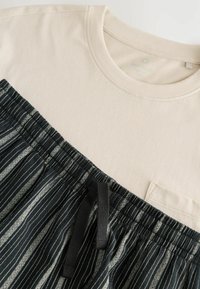 Helles beiges T-Shirt mit einem dunklen, gemusterten Stoffeinsatz am unteren Ende, der vertikale Streifen zeigt und über einen Kordelzug verfügt.