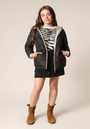 Zwarte zip-up jack met een metallic grijze voering, voorzien van een kleurgeblokte rits, gedragen over een zebra gestreept sweater en een zwarte rok. Bruine enkellaarsjes.