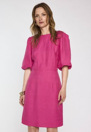 Woman with loose brown hair wearing a bright pink knee-length dress with puffed sleeves and gold bracelets on her left wrist.