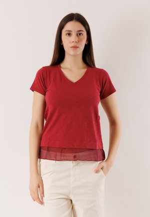 Donna con capelli castani lunghi, indossa una maglia rossa a maniche corte con scollo a V e orlo trasparente, e pantaloni beige chiaro, in piedi davanti a uno sfondo neutro.