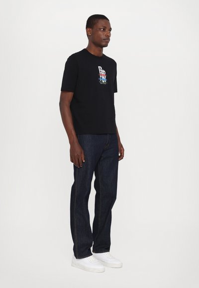 Czarny bawełniany t-shirt z wielokolorowym wzorem graficznym, w połączeniu z ciemnoniebieskimi dżinsami i białymi trampkami. Mężczyzna stoi na jednolitym tle.