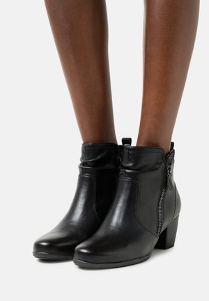 Botines de piel negra con un tacón bajo, textura lisa, cierre de cremallera lateral y detalles fruncidos en el tobillo.