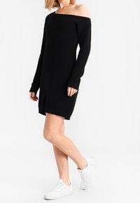 Schwarzes, schulterfreies Strickkleid mit gerippter Textur, langen Ärmeln und lockerem Schnitt. Getragen mit weißen Sneakern.