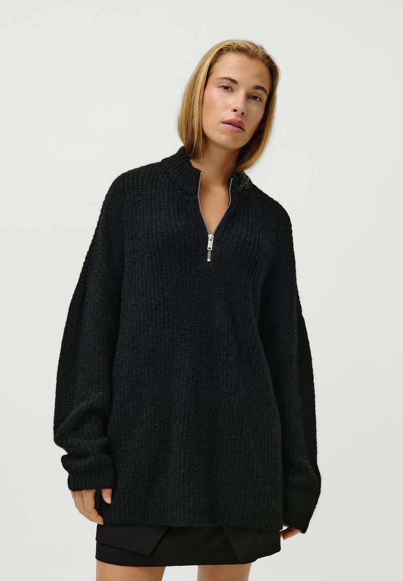 Kvinne iført en løs svart strikkegenser med en sølv half-zip og et svart miniskjørt, stående mot en enkel hvit bakgrunn.