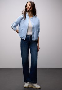 Mujer con chaqueta bomber azul claro, camiseta blanca, vaqueros anchos azul oscuro y zapatillas blancas, de pie contra un fondo gris.