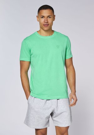 Man standing wearing a light green short-sleeve t-shirt and light gray shorts against a plain background.