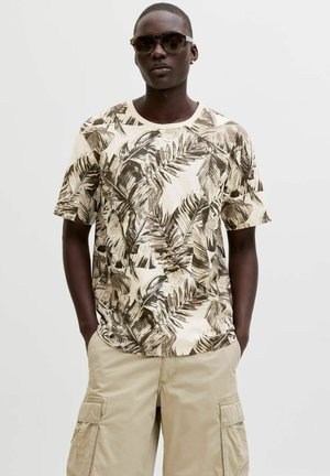 Man wearing brown and beige leaf-patterned t-shirt, beige cargo shorts, dark sunglasses, standing with hands in pockets on plain background.