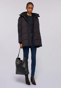 Černá péřová bunda s kapucí, prošívaný design, knoflíkové detaily, doplněná tmavými džínami a černou koženou kabelkou. Stylové kotníkové boty dokončují vzhled.
