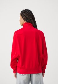 Giacca pullover rossa con collo alto, maniche raglan, tessuto testurizzato e polsini a coste. Presenta una vestibilità ampia e orlo elastico.