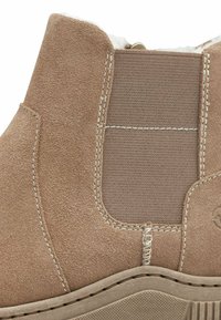 Bottine en daim beige avec un panneau en tissu, des coutures contrastées et une semelle en caoutchouc texturée. Les accents comprennent une petite fermeture éclair en métal.