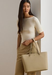 Ženska z dolgimi temnimi lasmi, oblečena v kremno majico in bež hlače, drži veliko bež usnjeno torbo z zlatimi dodatki.