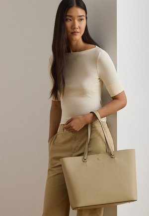 Kobieta z długimi ciemnymi włosami, ubrana w kremowy top i beżowe spodnie, trzymająca dużą beżową skórzaną torbę na ramię ze złotymi akcentami.