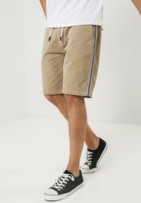 Shorts beiges avec des détails latéraux rayés noirs et blancs, fabriqués en mélange de coton, dotés d'une taille élastique avec cordons de serrage. Baskets noires.