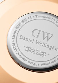 Tył zegarka Daniel Wellington, wykonany ze stali nierdzewnej w kolorze srebrnym, z wygrawerowanym logo, numerem modelu i numerem seryjnym, otoczony różowym złotym kopertą.