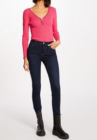 Haut rose côtelé à manches longues avec un col en V et un détail zippé, associé à un jean slim bleu foncé et des bottines noires.