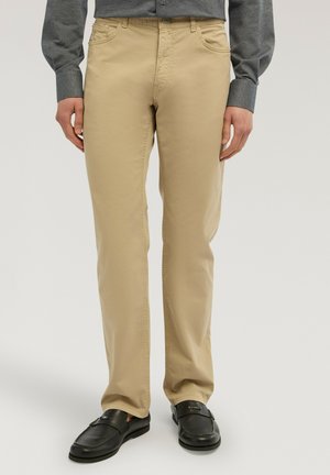 Uomo che indossa pantaloni beige a gamba dritta, camicia grigia a maniche lunghe e mocassini neri in pelle, in piedi con le mani rilassate ai lati.