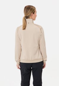 Beige softshell jack met een hoge kraag, geribde manchetten en een gladde textuur. Beschikt over contrasterende schouderpanelen en een aansluitend ontwerp.