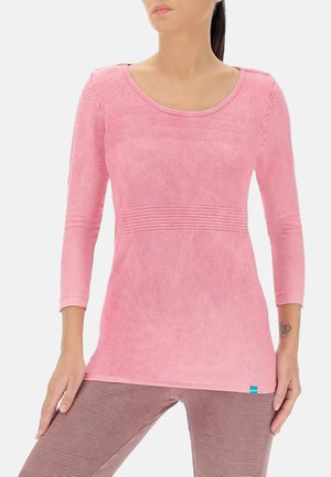 Woman wearing long-sleeve pink textured top and mauve ribbed leggings, standing with one hand on hip and the other resting on thigh.
