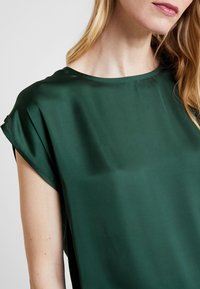 Grøn satin top med en afslappet halsudskæring og korte, opsmøgede ærmer. Har en glat tekstur og flydende design. Nærbillede.