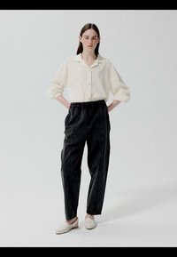 Kobieta stojąca z rękami w kieszeniach czarnych spodni, ubrana w białą koszulę na guziki i białe buty wsuwane na neutralnym tle.