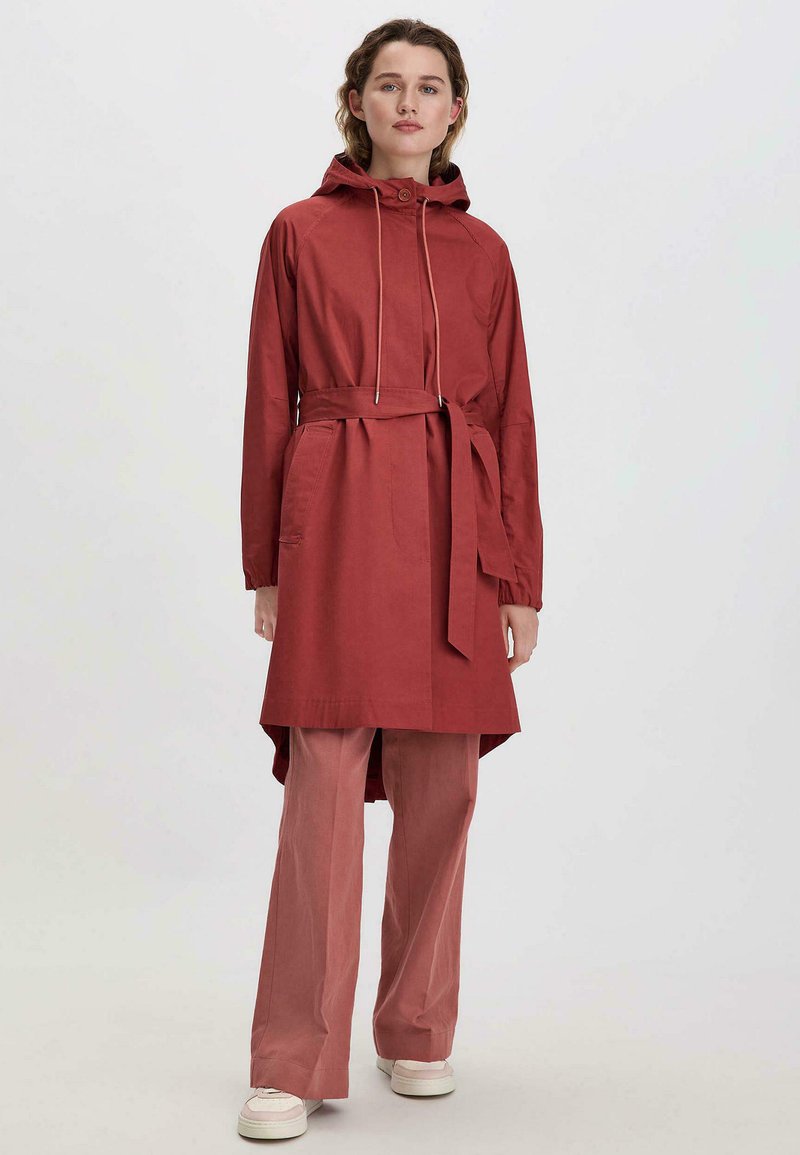 Rote Baumwolljacke mit Kapuze, Kordelzug und Gürtel. Mit langen Ärmeln,Seitentaschen und einem längeren hinteren Saum ausgestattet. Über passenden Hosen getragen.