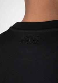 Černé tričko s kulatým výstřihem a jemně vyšitým logem "WRONG FRIENDS" na zadní straně. Hladká textura a přiléhavý střih.