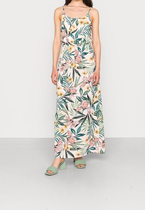 Frau trägt ein ärmelloses weißes Maxikleid mit pinkem und gelbem Blumenmuster und grüne Absatzsandalen mit geflochtenen Riemen.