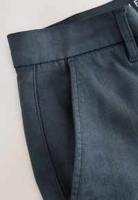 Gros plan sur la ceinture et la poche latérale d’un pantalon bleu marine avec des coutures visibles sur un fond clair.