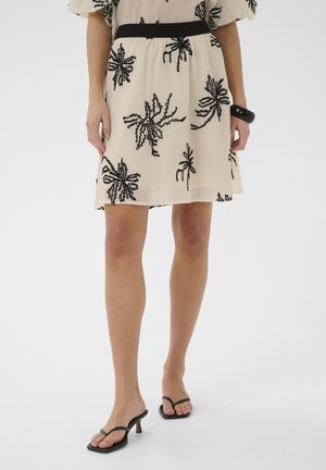 Woman wearing a beige skirt with black floral patterns, black belt, black bracelet, and black heeled sandals standing against a white background.