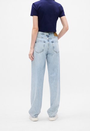 Personne portant un jean bleu clair ample et un t-shirt court bleu marine, debout de dos face à la caméra sur fond blanc.
