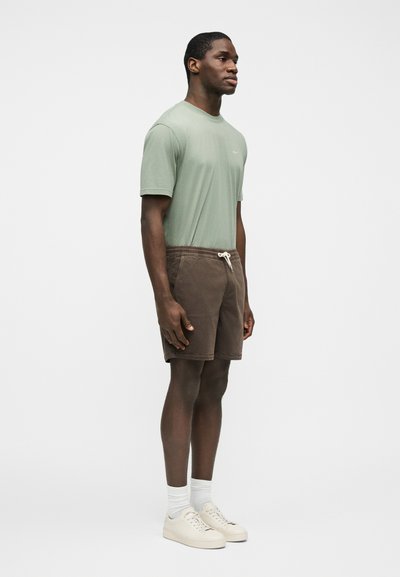 Homme debout portant un t-shirt vert clair, un short marron à cordon, des chaussettes blanches et des baskets beige sur un fond uni.