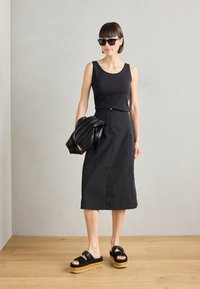 Svart tank topp, knee-length svart skjørt med frontknapper, kraftige svarte sandaler med korksåler, holder en svart teksturert omsorg.