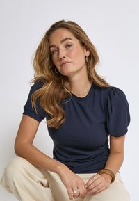 Marinblus med ribbat mönster, korta puffärmar och rund halsringning, tillverkad av ett mjukt material. Modellen bär ljusa byxor och accessoarer.