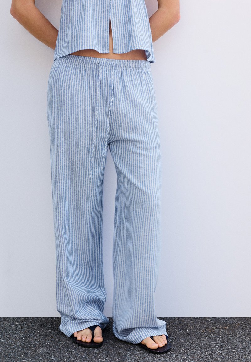 Personne portant un pantalon ample à rayures verticales bleues et blanches avec un top court assorti, debout pieds nus sur un trottoir sombre contre un mur blanc.