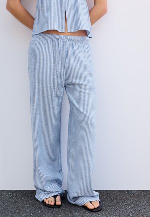 Persona che indossa pantaloni larghi a righe verticali blu e bianche con top corto abbinato, in piedi a piedi nudi su pavimento scuro contro parete bianca.
