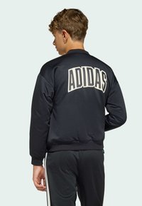 Svart träningsjacka med ribbade ärmslut och midja, med en stor vit "ADIDAS"-logotyp på ryggen i fet stil.