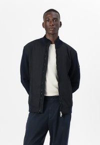 Μαύρο μπουφάν με ψηλό γιακά, μπροστινό φερμουάρ και ραμμένα μανίκια; σε συνδυασμό με ανοιχτόχρωμο μπλουζάκι και ναυτικά παντελόνια.