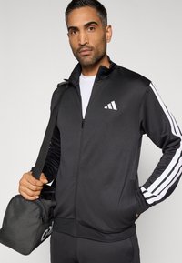 Čierna športová bunda s bielymi tromi pruhmi, so zapínaním na zips a vysokým golierom; doplnená o čiernu tašku s logom.