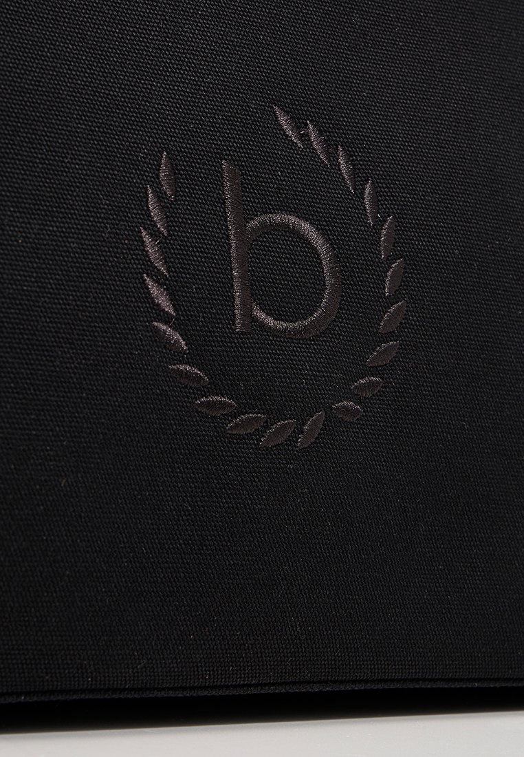 Lettera minuscola "b" ricamata in filo scuro, circondata da un ramo di alloro su un tessuto nero testurizzato.