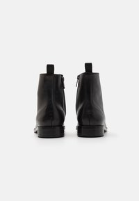 Botas de cuero negro hasta el tobillo con un acabado liso, que cuentan con cremalleras laterales y tiradores. La suela es plana y ligeramente texturizada.