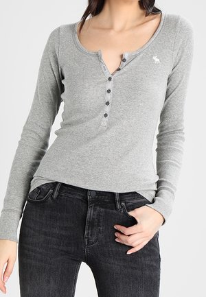 Vrouw draagt een aansluitende grijze henley top met lange mouwen en zwarte knopen, gecombineerd met zwarte high-waisted jeans, hand in de zak.