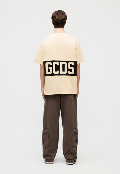 Άτομο που φοράει μπεζ φαρδιά μπλούζα με έντονο μαύρο κείμενο "GCDS" στην πλάτη, συνδυασμένη με χαλαρό καφέ παντελόνι cargo και αθλητικά παπούτσια.