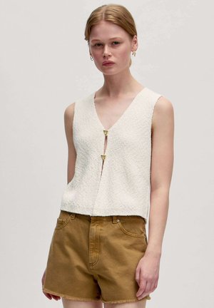 Witte textuurloze mouwloze top met een V-hals en twee gouden driehoekige sluitingen, gecombineerd met bruine denim shorts.